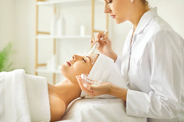 Woman enjoys professional skin care at spa beauty salon. Side view beautician applying cleansing moisturizing facial mask on face of young woman lying on bed in white towel. Facial treatment concept